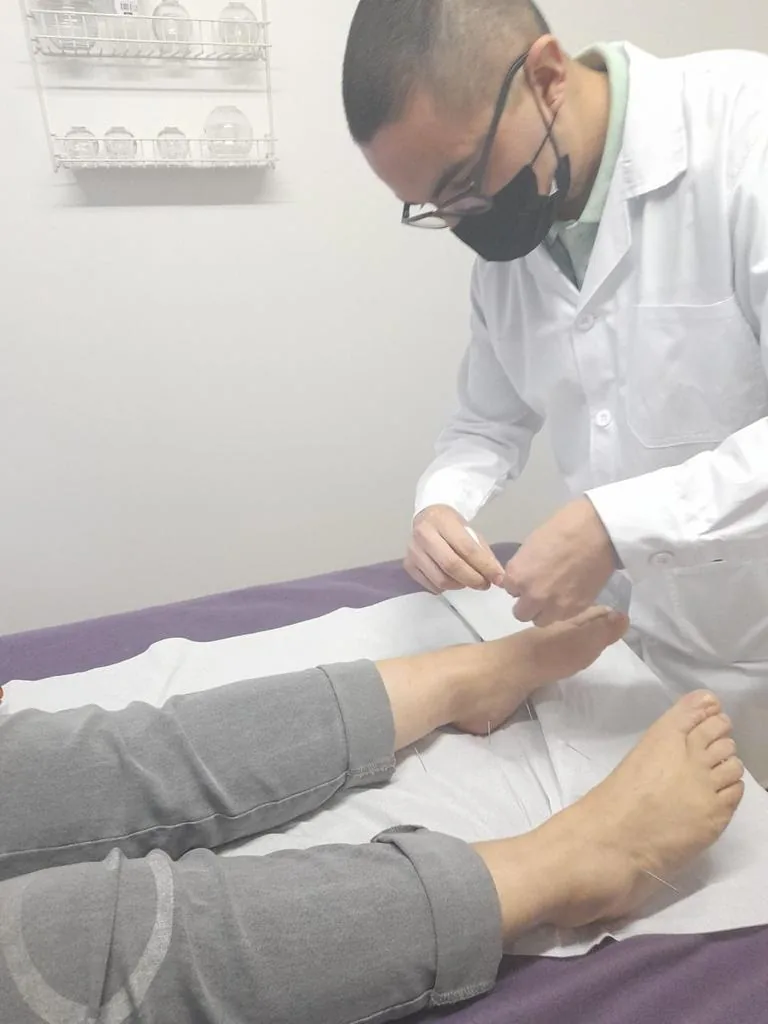 Alumno aplicando agujas de acupuntura en los puntos de los pies de un paciente.