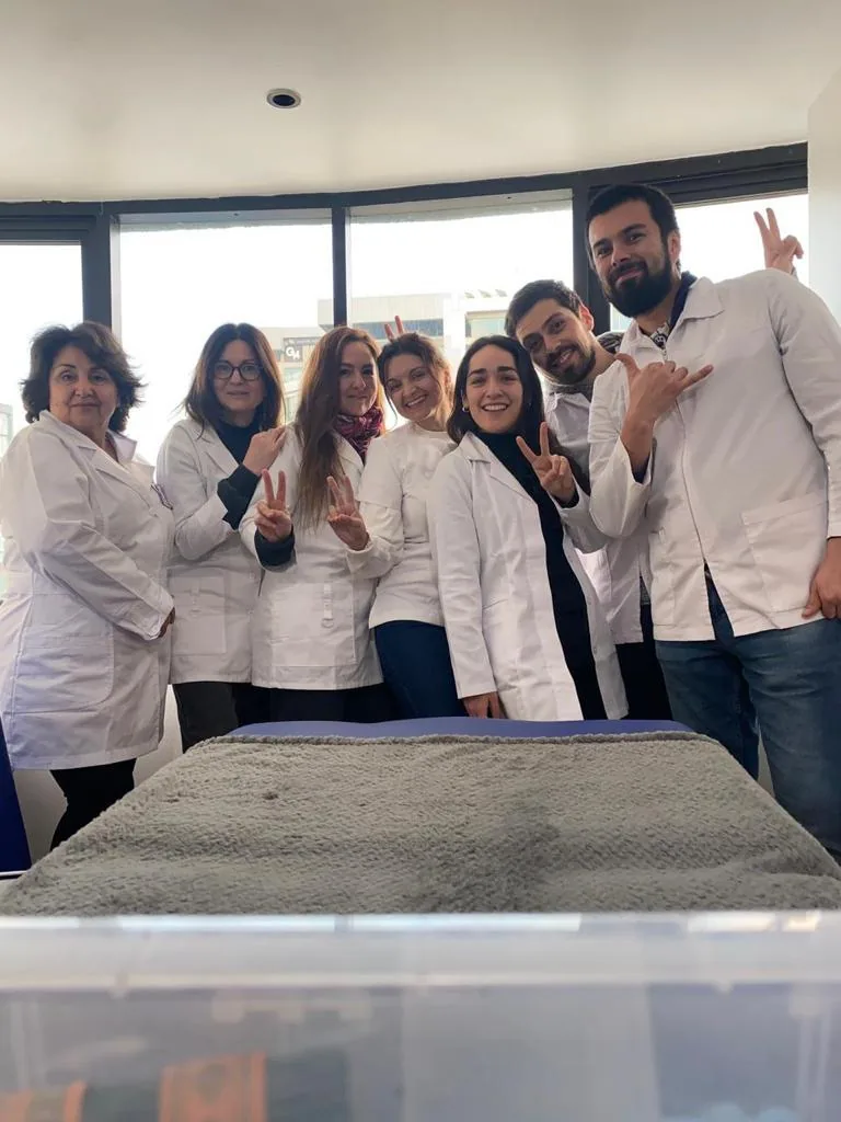 Grupo de alumnos y tutores en bata clínica, junto a una camilla de práctica en una de las sedes.