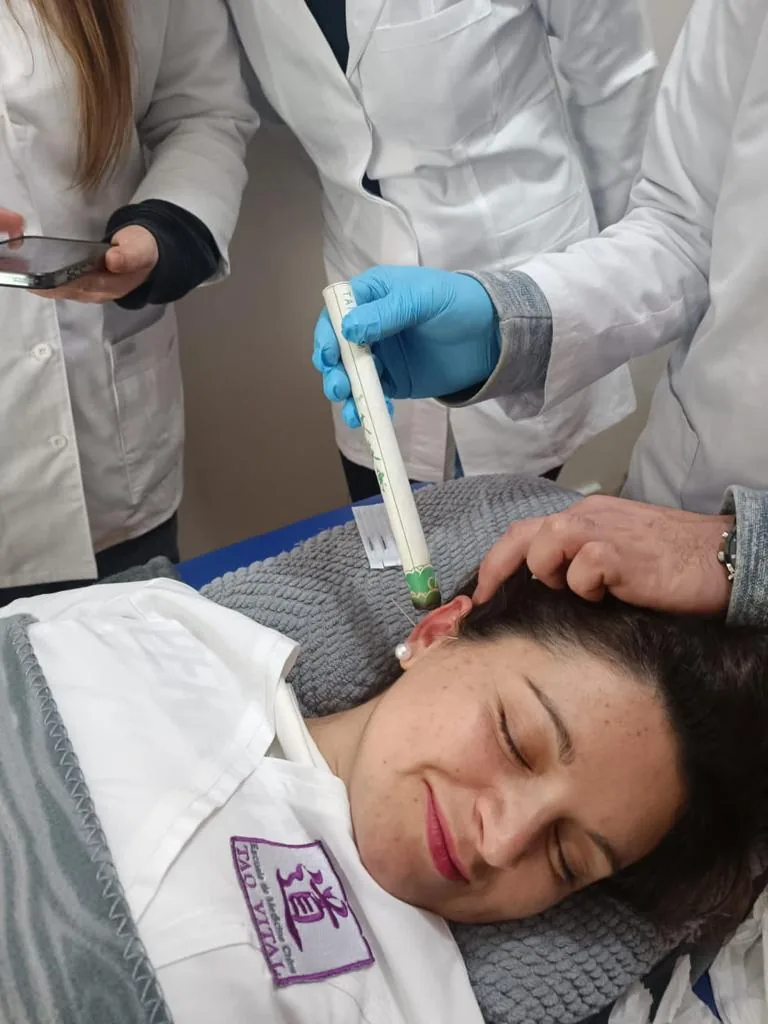 Docente colocando semillas de auriculoterapia con pinzas en la oreja de una paciente.